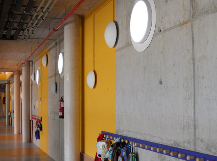 Couloir d'école avec éclairage encastré et murs jaunes. La lumière naturelle pénètre par des fenêtres circulaires.