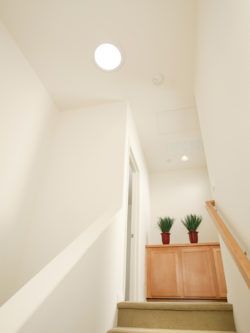 Conduit de lumière circulaire au plafond, escalier blanc et meuble en bois.